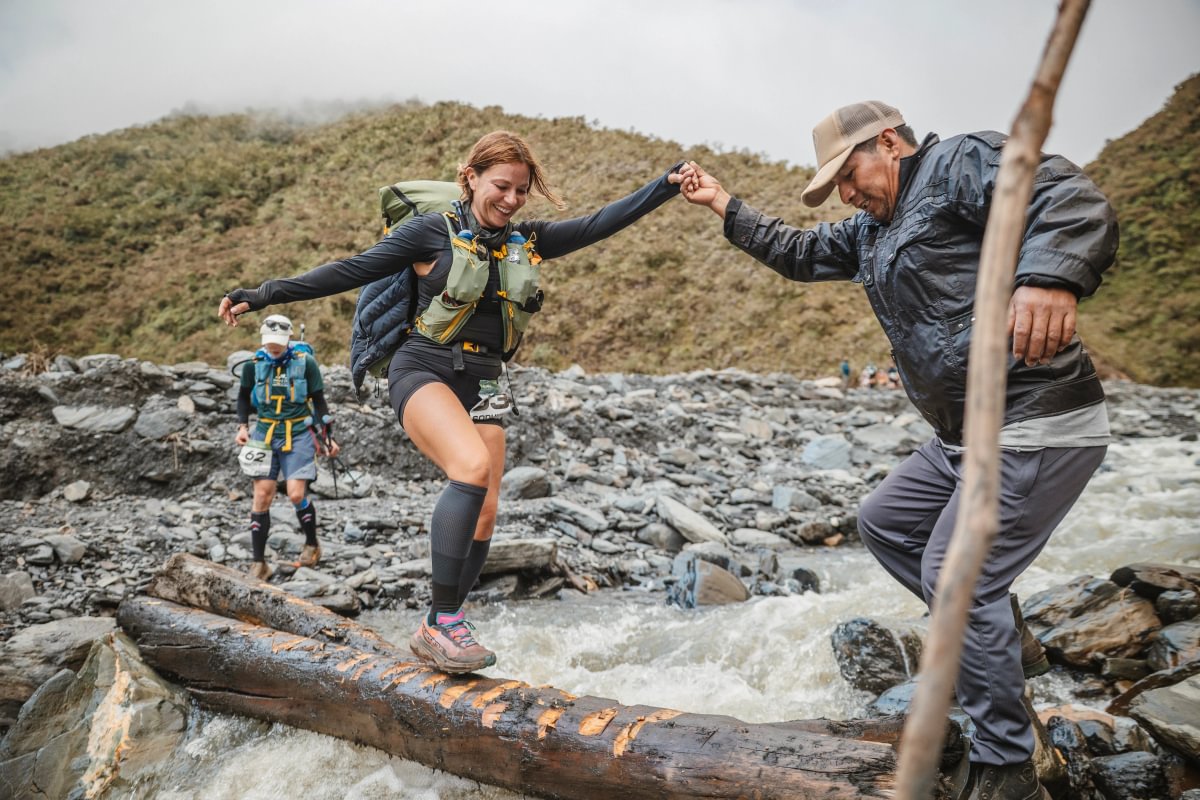 A runner crossing a log bridge over a mountain river with the help of a local guide during the BTU Jungle Ultra, Peru
